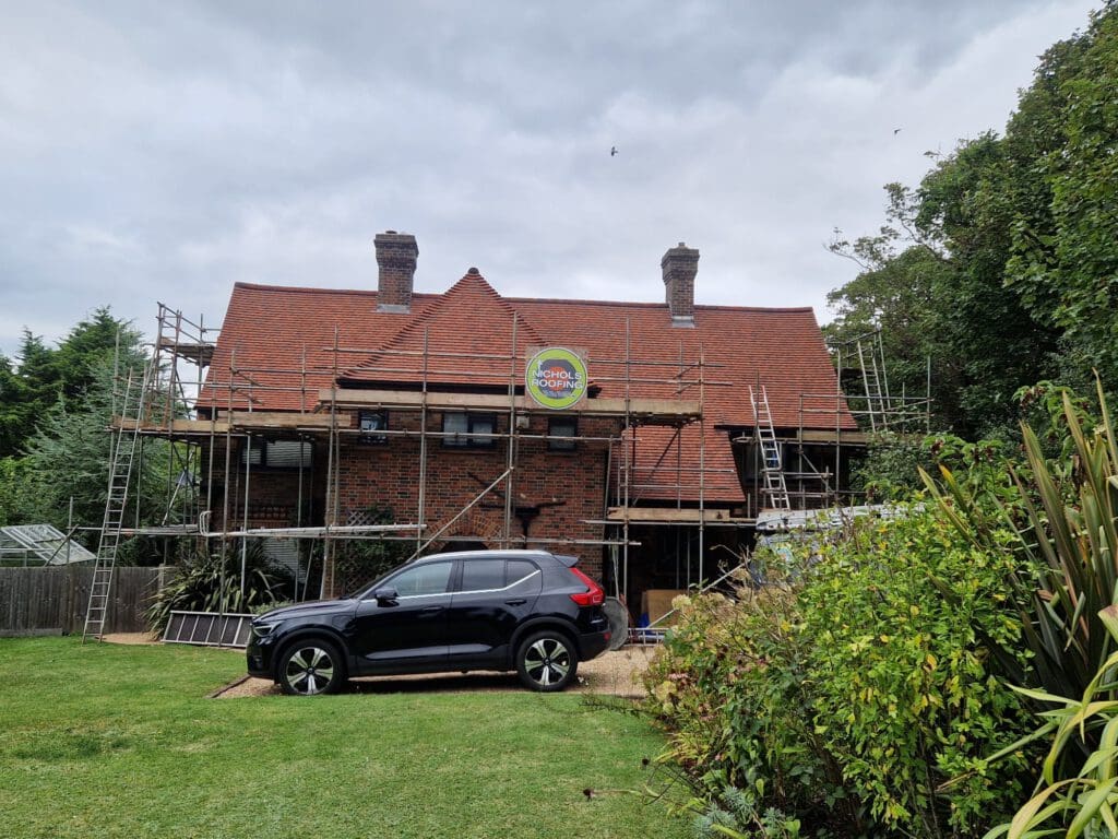 A red-brick house with a red-tiled roof is under renovation. Scaffolding surrounds the building, and a black car is parked in front of it. A circular sign is attached to the house, and the scene is set in a lush garden.