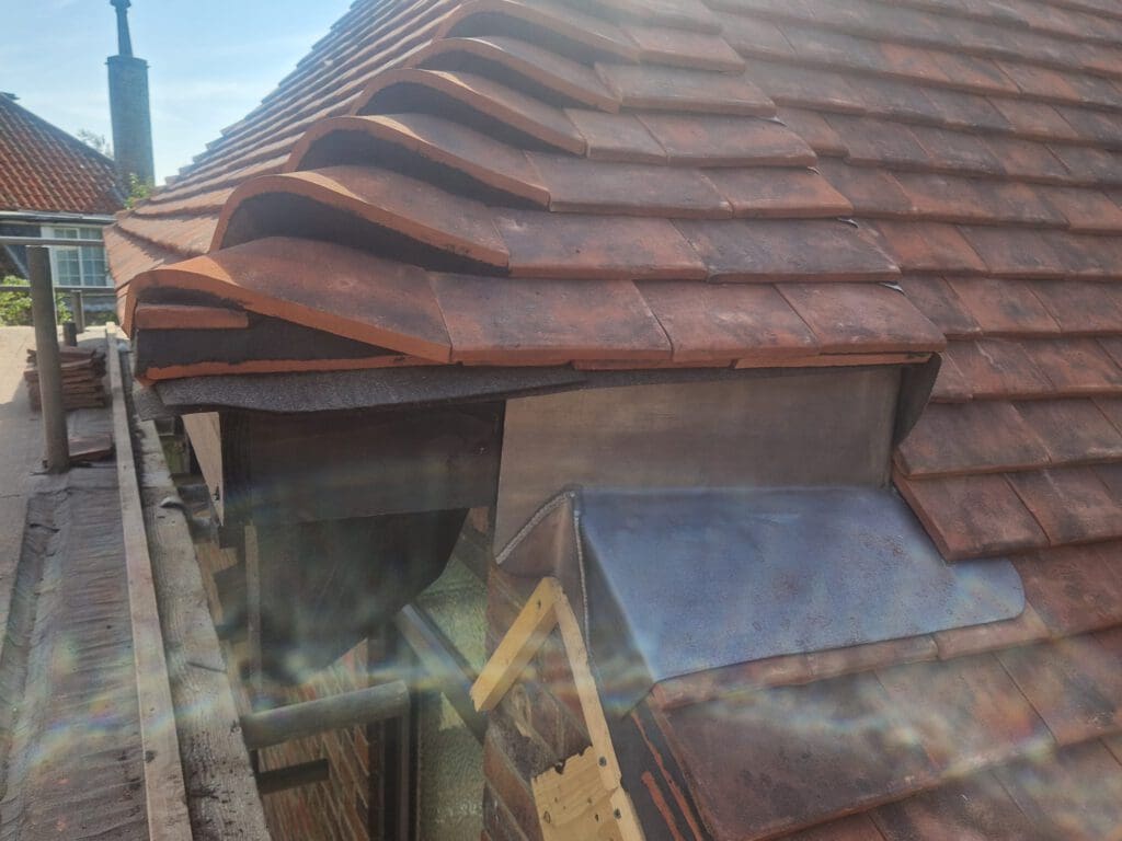 Close-up of a red tiled roof with overlapping tiles near the edge. A small metal vent is visible beneath the tiles. Sunlight creates a lens flare effect, and a portion of the scaffold and neighboring roof can be seen.