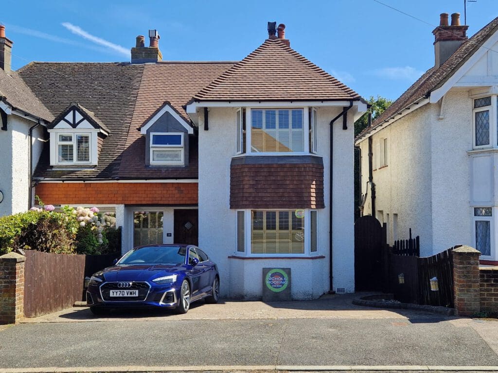 A suburban house with a brick chimney and brown roof features a double door entrance. A blue car is parked in the driveway. The house is flanked by two similar houses, under a clear blue sky.