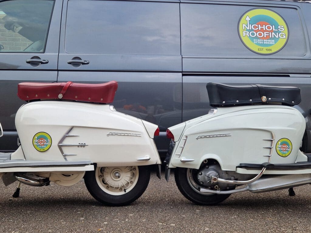 Two vintage scooters with red and black seats are parked side by side in front of a gray van. Both scooters and the van display circular "Nichols Roofing" logos.
