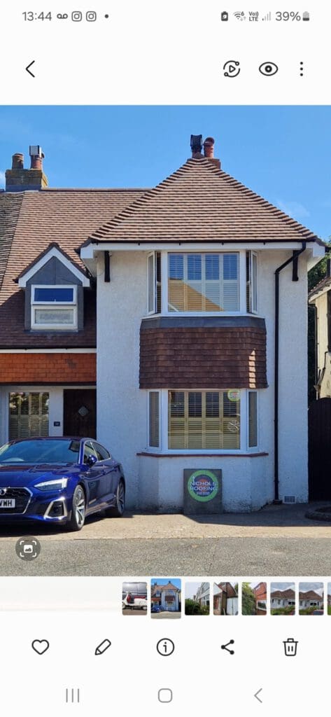 A two-story house with a white exterior and reddish-brown roof, featuring a large bay window. There is a blue car parked in the driveway, and a round sign displayed on the front wall.