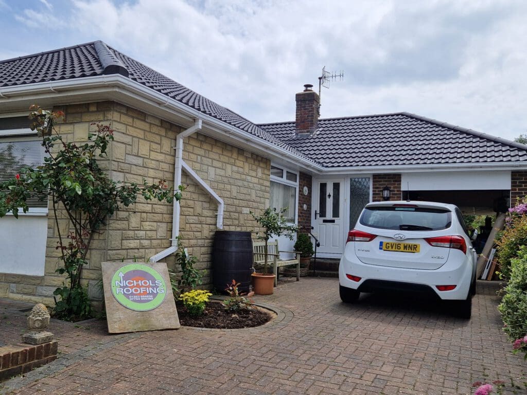 A single-story house with a tiled roof and a driveway. A white car is parked in the driveway. A "Nichols Roofing" sign leans against the house. There's a small garden with plants near the entrance.