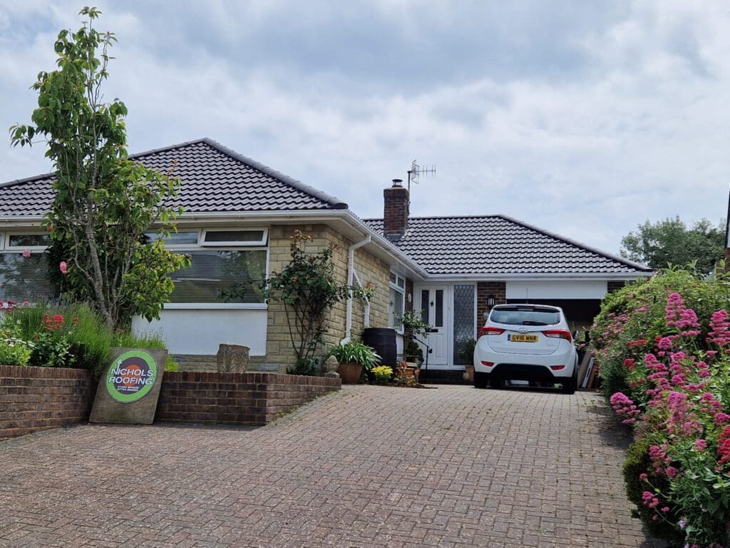 A brick bungalow with a tiled roof, surrounded by greenery and colorful flowers. A white car is parked in the driveway. A sign reads "Nichols Roofing." The sky is overcast.