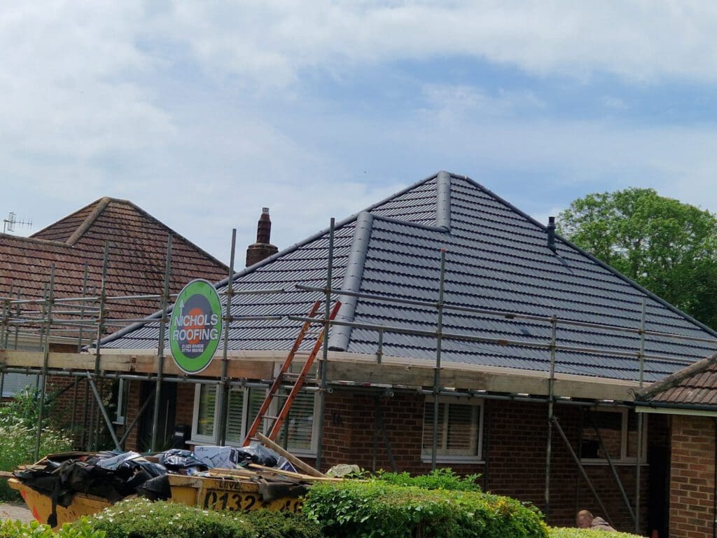 A newly renovated roof on a brick house is surrounded by scaffolding. A sign reads "Nicholls Roofing." A skip filled with debris is in front of the house, and trees can be seen in the background under a partly cloudy sky.