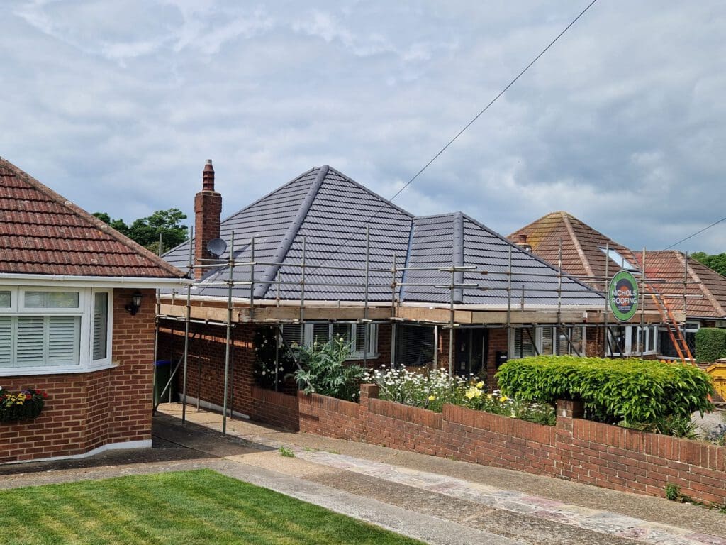 A brick house with a newly tiled gray roof is surrounded by scaffolding. The front yard features a lush green lawn and blooming flowers. Overcast sky in the background.