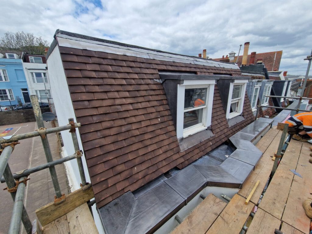 A construction worker is on scaffolding beside a sloped roof of a residential building. The roof is covered with brown shingles and features two dormer windows. The sky is partly cloudy.