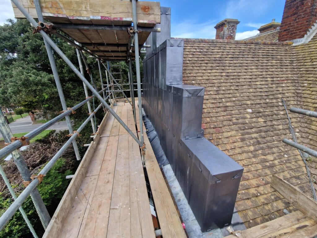 Scaffolding surrounds the side of a building with a steep, shingled roof. Metal cladding covers a section of the wall, partially obscured by the wooden scaffolding. Trees are visible in the background under a clear blue sky.