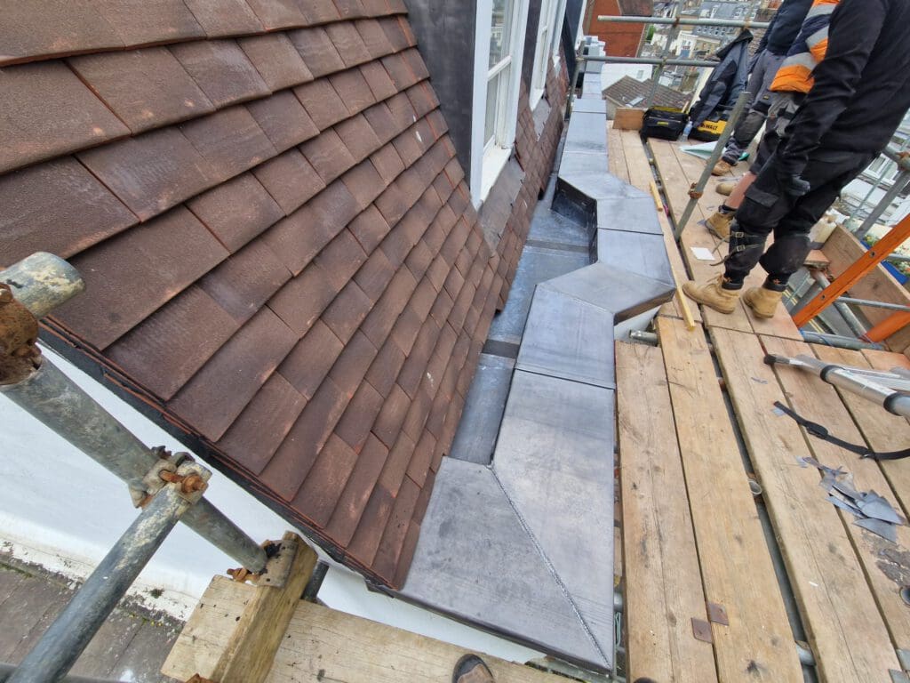 Workers standing on scaffolding next to a building's roof. The roof has brown shingles and new metal flashing is being installed. Construction tools and materials are visible on the wooden platform.