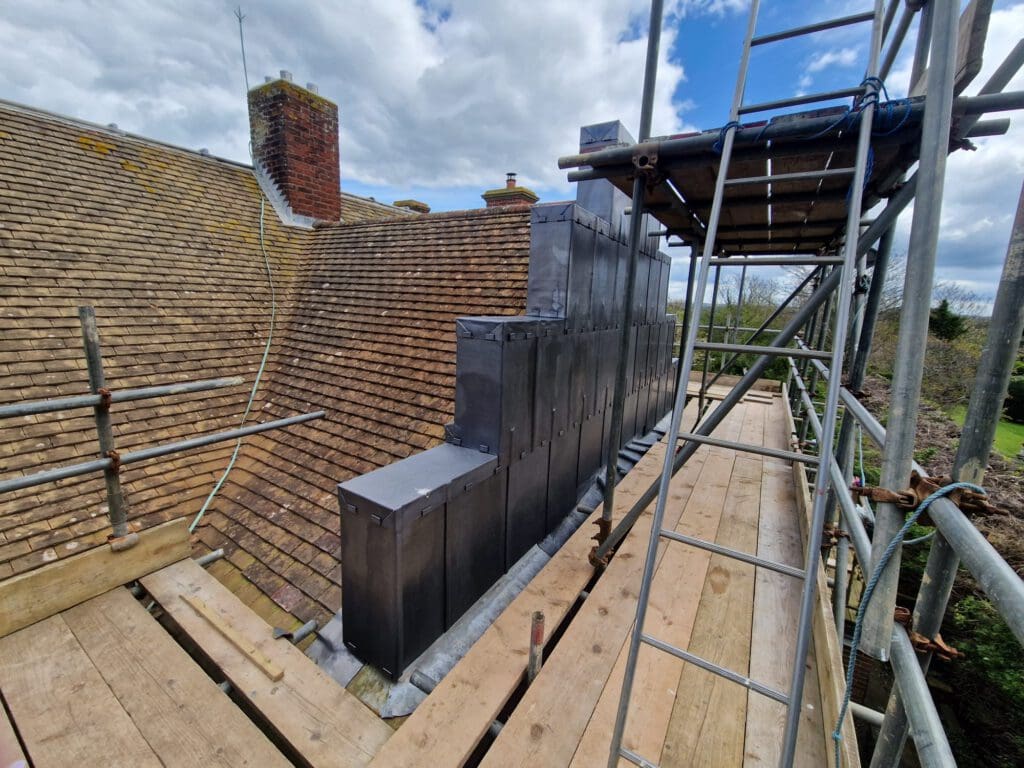 Scaffolding on a rooftop beside a chimney, with recently installed lead covering. Cloudy sky overhead and trees visible in the background. Building materials and wooden planks are part of the construction setup.