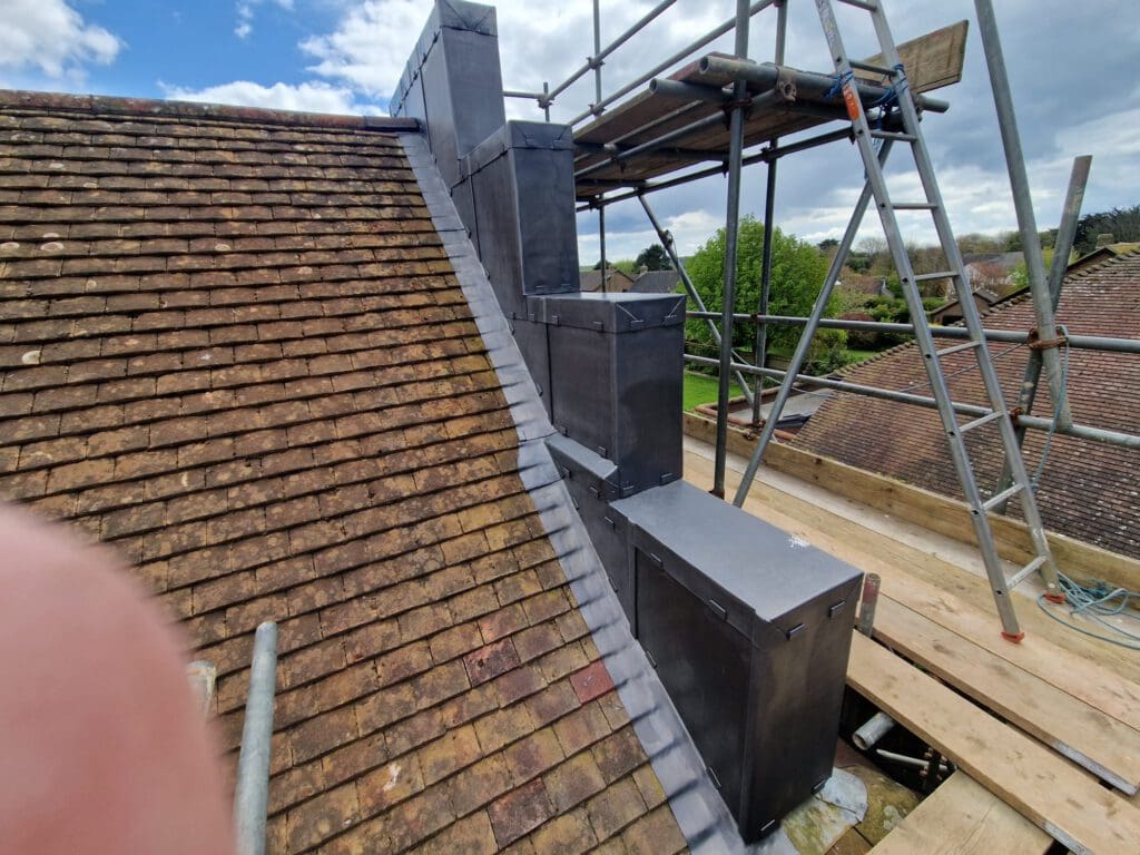 Roof construction showing scaffolding on the right and newly installed grey metal cladding around a chimney. The roof has brown shingles and a background of a cloudy sky and trees. A thumb partially covers the lens in the bottom left corner.