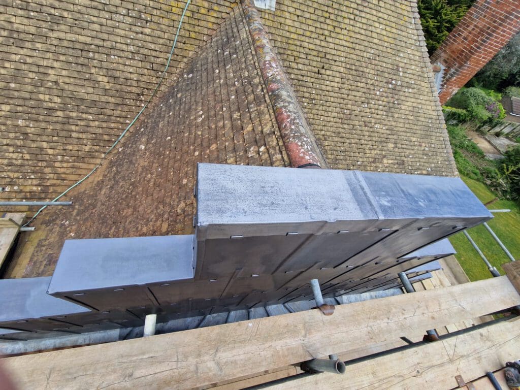 View from above of a steeply pitched roof with weathered shingles. A section is under renovation with new, shiny metal cladding. Scaffolding is visible in the foreground, with a chimney and trees in the background.