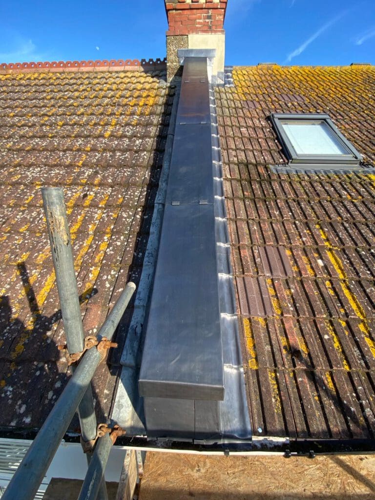 A close-up view of a rooftop with discolored tiles and a newly installed lead chimney cover. There's visible scaffolding on the left, and a skylight window is set into the roof on the right. The sky is clear and blue.