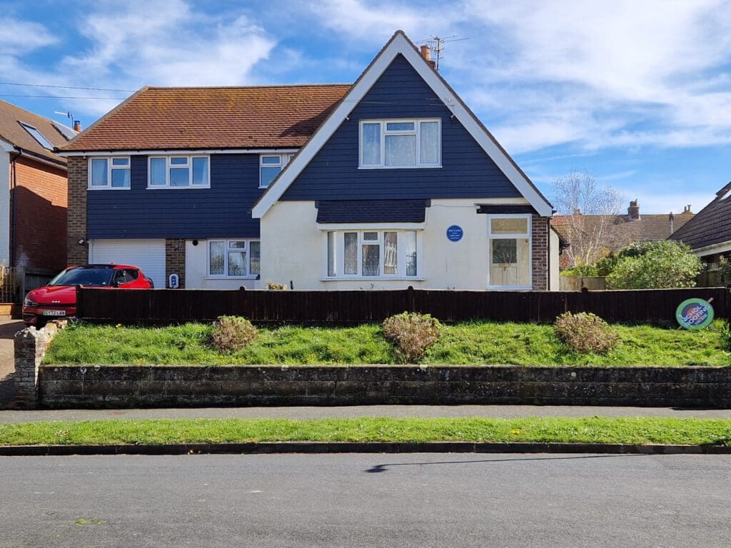 A two-story house with a triangular roof and blue upper facade. It features a well-kept lawn with shrubs, a driveway with a red car, and a visible round blue plaque on the house. The sky above is clear with some clouds.