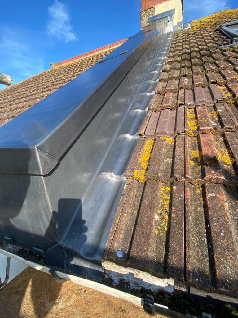 A sloped rooftop with weathered, moss-covered tiles. A newly installed metal ridge is visible, shining under a clear blue sky. The contrasting textures highlight the difference between the old and new roofing materials.