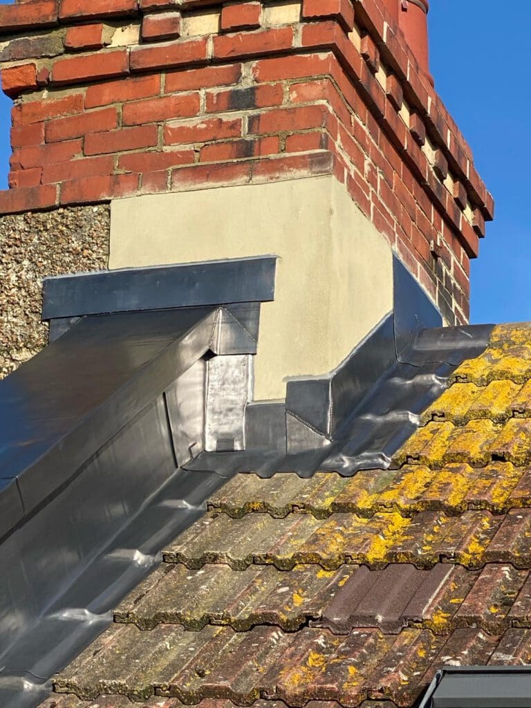 A close-up of a brick chimney with metal flashing on a tiled roof. The tiles are weathered with moss and the sky is clear and blue.