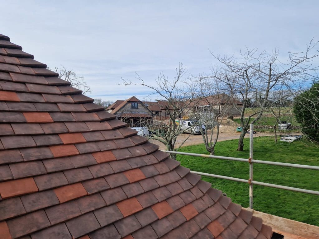 View from a rooftop shows a curved brown shingle roof in the foreground. Below, there is a green lawn with bare trees and construction materials. In the background, several houses are visible under a cloudy sky.