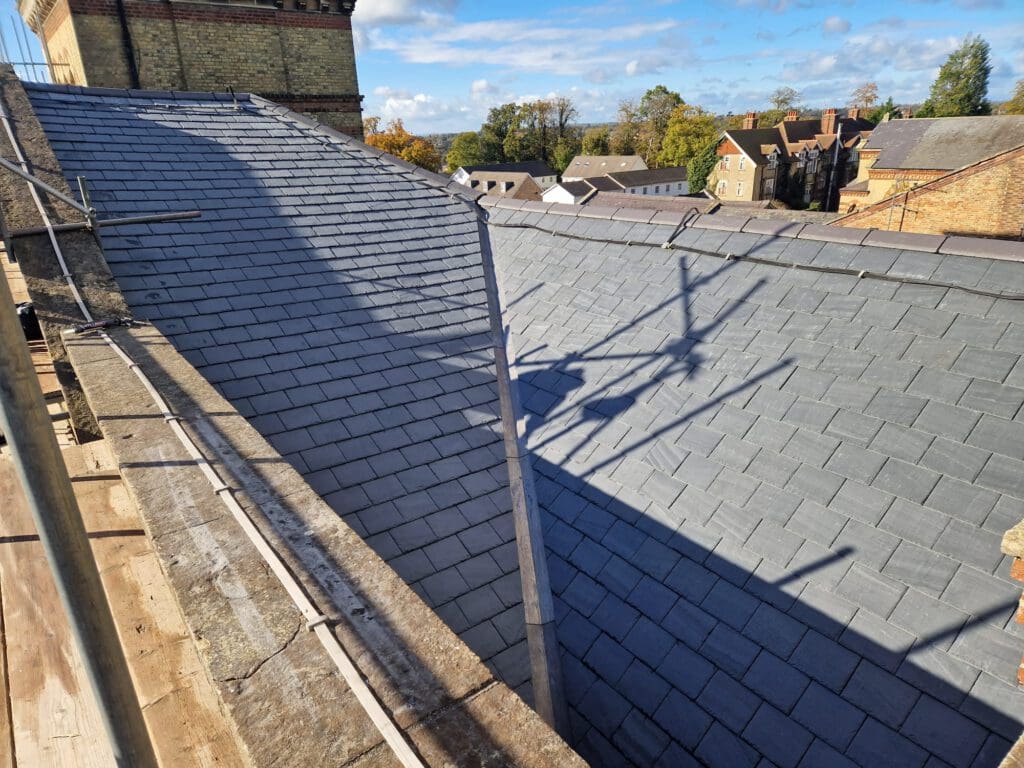 View from a rooftop, showing angled grey slate tiles with clear blue sky above. Scaffolding casts shadows on the roof. Surrounding buildings and trees are visible in the background.