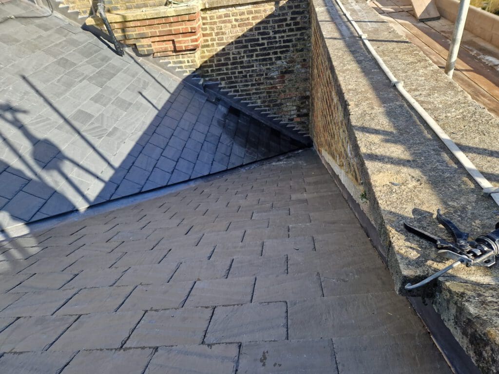 A rooftop with newly installed brown shingles, adjacent to a brick wall. Shadows of a ladder and other objects are visible. A metal tool is placed on the upper right corner of the roof.