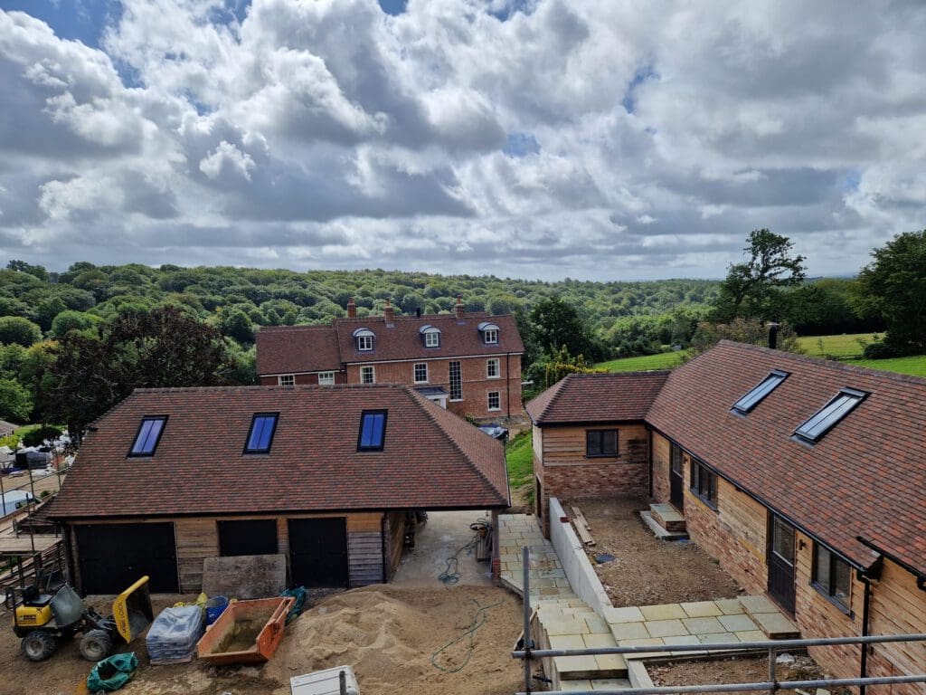 A scenic view of countryside buildings under construction. Two houses with red-tiled roofs are in the foreground, with construction materials visible. A large brick house and lush green hills are in the background under a partly cloudy sky.
