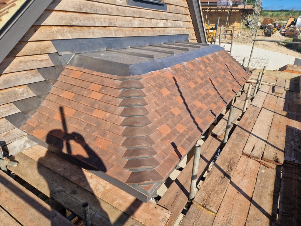 A newly constructed sloped roof with reddish-brown tiles and metal flashing is surrounded by wooden scaffolding. A person's shadow holding a tool is visible on the roof. Nearby, more construction materials and work are in progress.