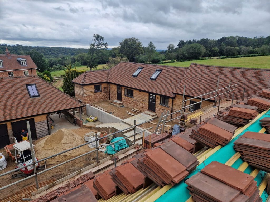 A construction site with several buildings featuring red tiled roofs. Scaffolding is set up, and bricks are stacked in the foreground. The surrounding area includes trees and green fields under a cloudy sky.