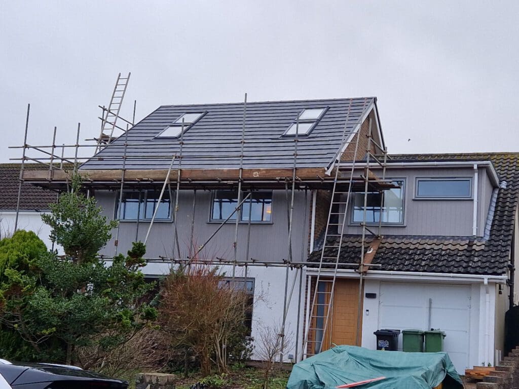A two-story house with gray siding is under renovation. Scaffolding surrounds the upper section, including a partially completed roof with gray tiles. Ladders lead to the roof. The yard has trees, and two trash bins are in front of the garage.