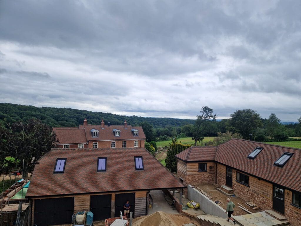 A large, brick country house with red-tiled roofs is surrounded by lush green trees and fields under a cloudy sky. Construction work is happening in the front yard, with workers visible near a large pile of sand.