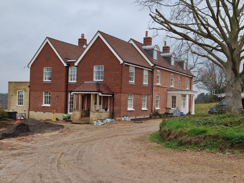 A large two-story brick house with a gabled roof is under construction. The building features multiple chimneys and several windows. The area around the house is unpaved with piles of construction materials and machinery visible. A large tree stands nearby.