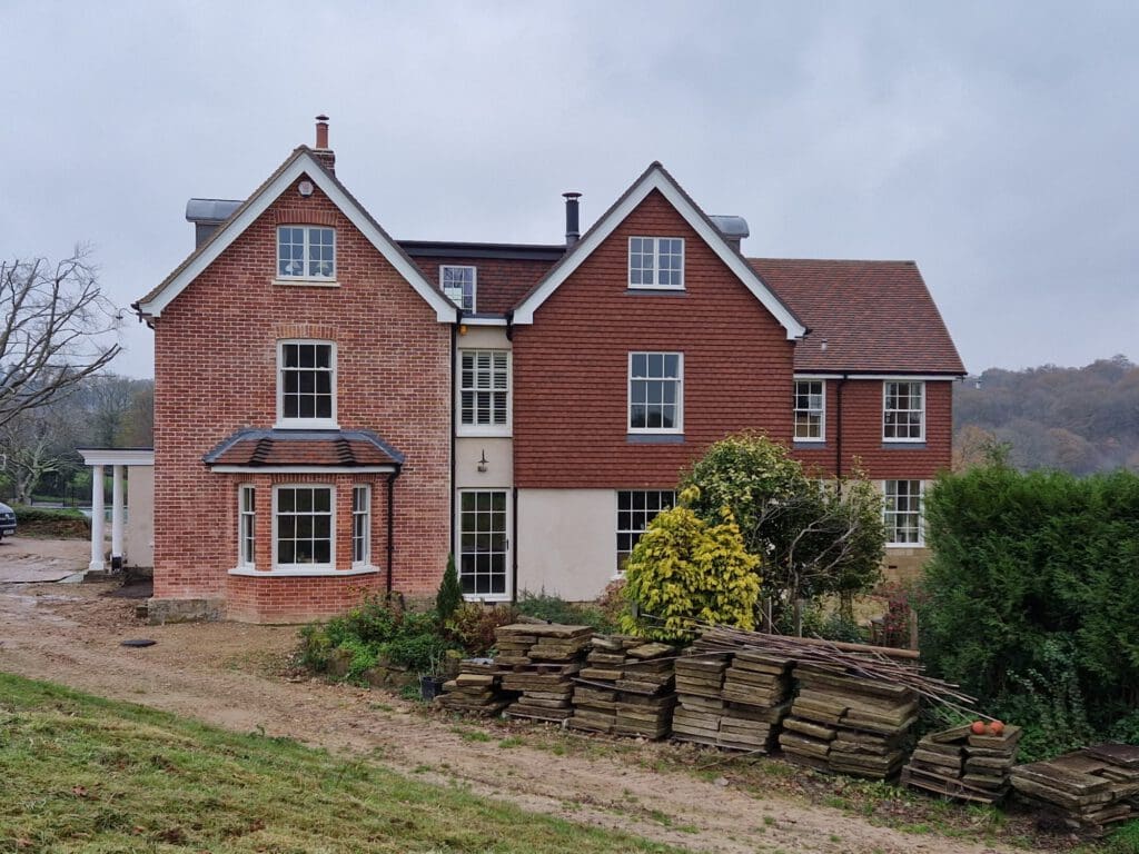 A large two-story red brick house with multiple gabled roofs sits on a grassy plot. Windows adorn the upper and lower floors. In front, there's a stack of wooden planks and shrubs alongside the building. The sky is overcast.