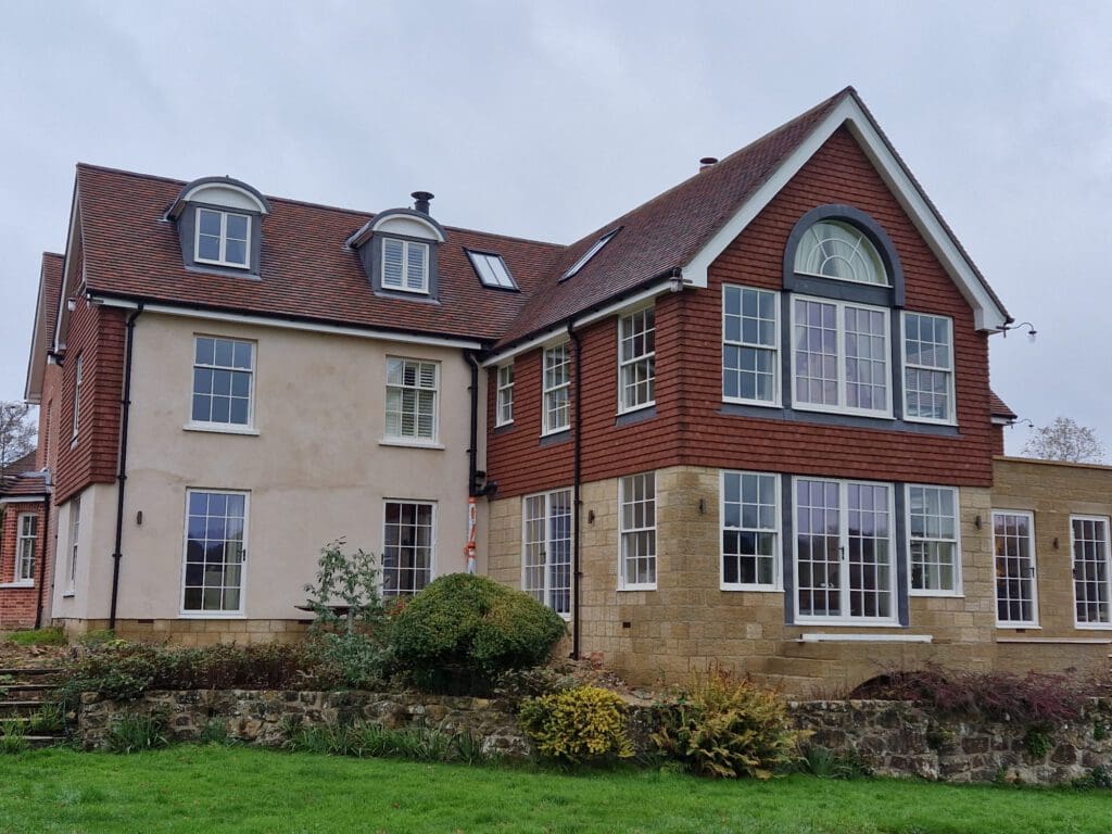 A large, multi-story house with a mix of beige and redbrick exterior walls, numerous windows, and a steep, dark red roof. The house is surrounded by a neatly landscaped garden with green grass and various shrubs.