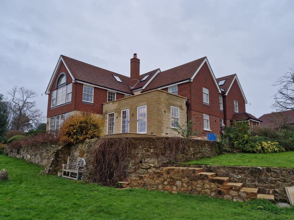 A large two-story red brick house with a chimney and multiple windows, featuring a stone patio extension. The house sits on a grassy hill with a stone retaining wall and a bench on the lawn. Overcast sky in the background.