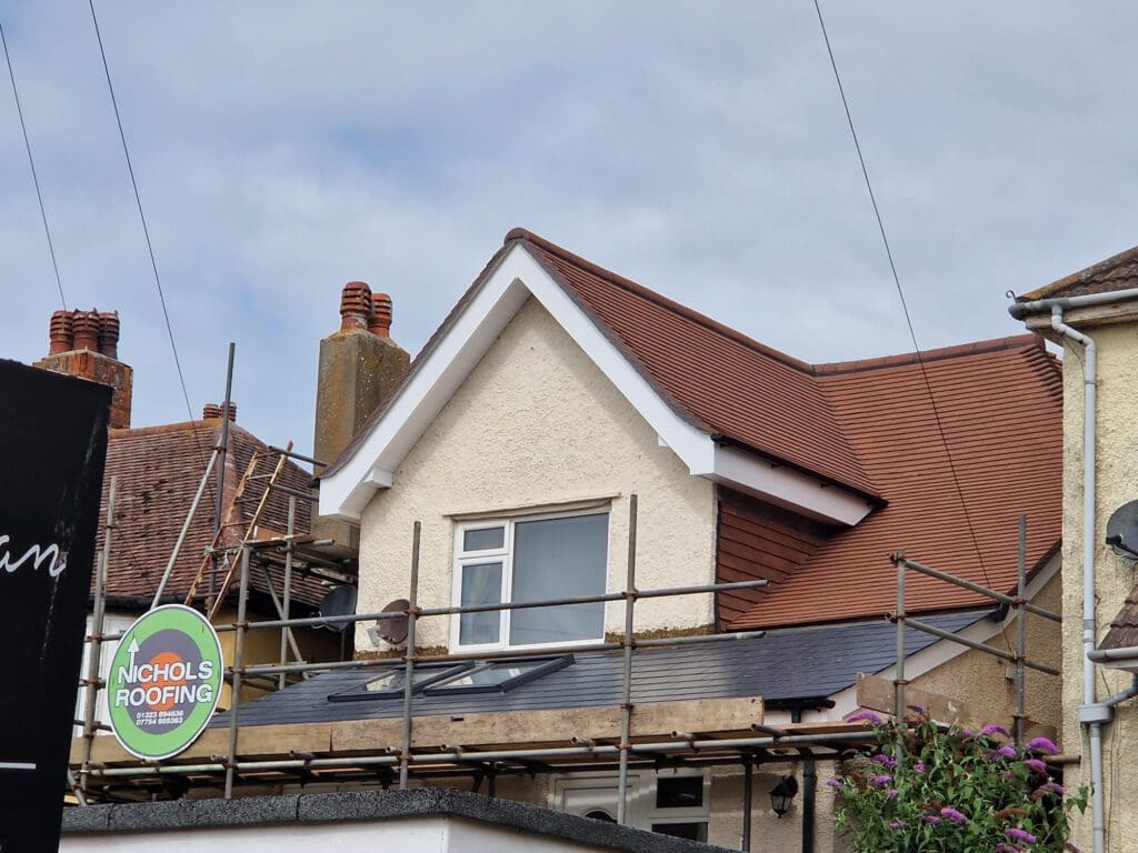 A house with beige walls and a brown shingled roof is under renovation. Scaffolding surrounds the front, and a sign reading "Nichols Roofing" is visible. The sky is cloudy in the background.