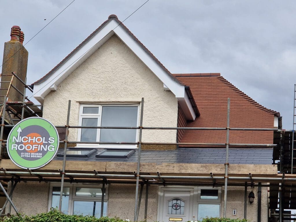 A house with beige walls is under renovation. The roof features red and grey tiles. Scaffolding surrounds the structure with a sign reading "Nichols Roofing" and phone numbers. A small window is visible below the roof. The sky is overcast.