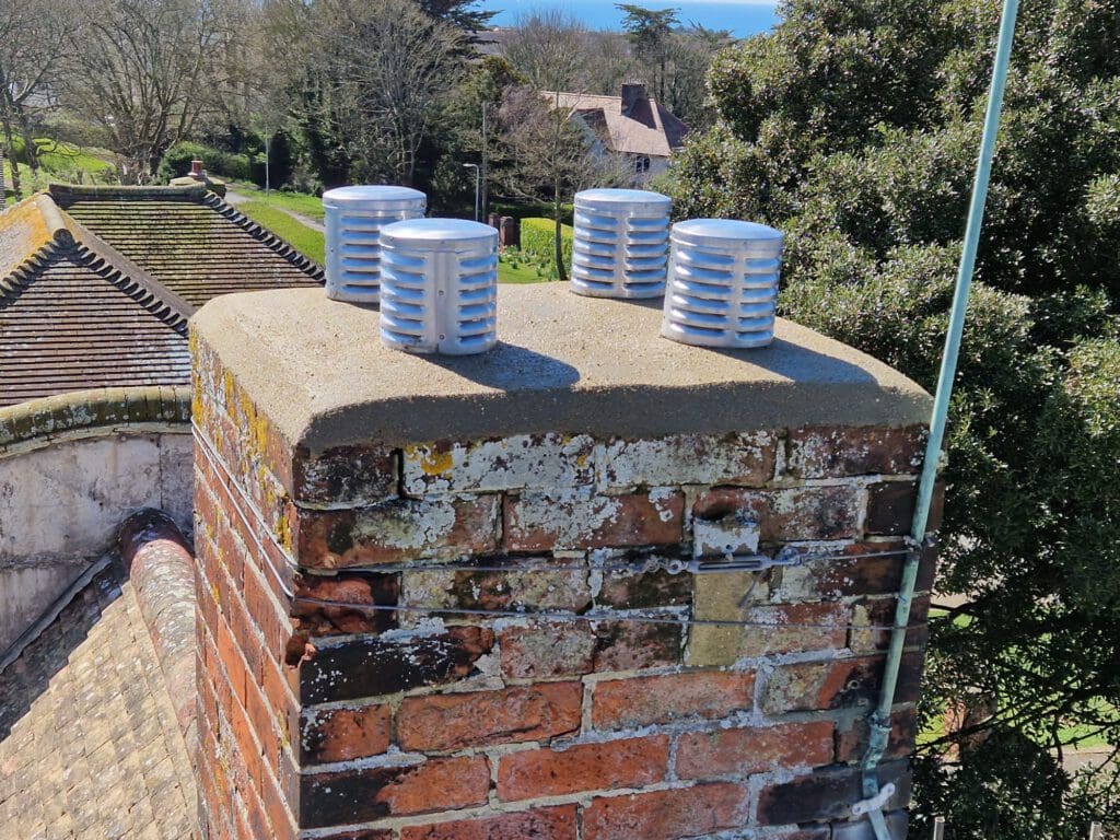 Brick chimney with four metal vent caps on top, set against a backdrop of trees and a house. The vents are cylindrical and silver, while the chimney shows signs of weathering.