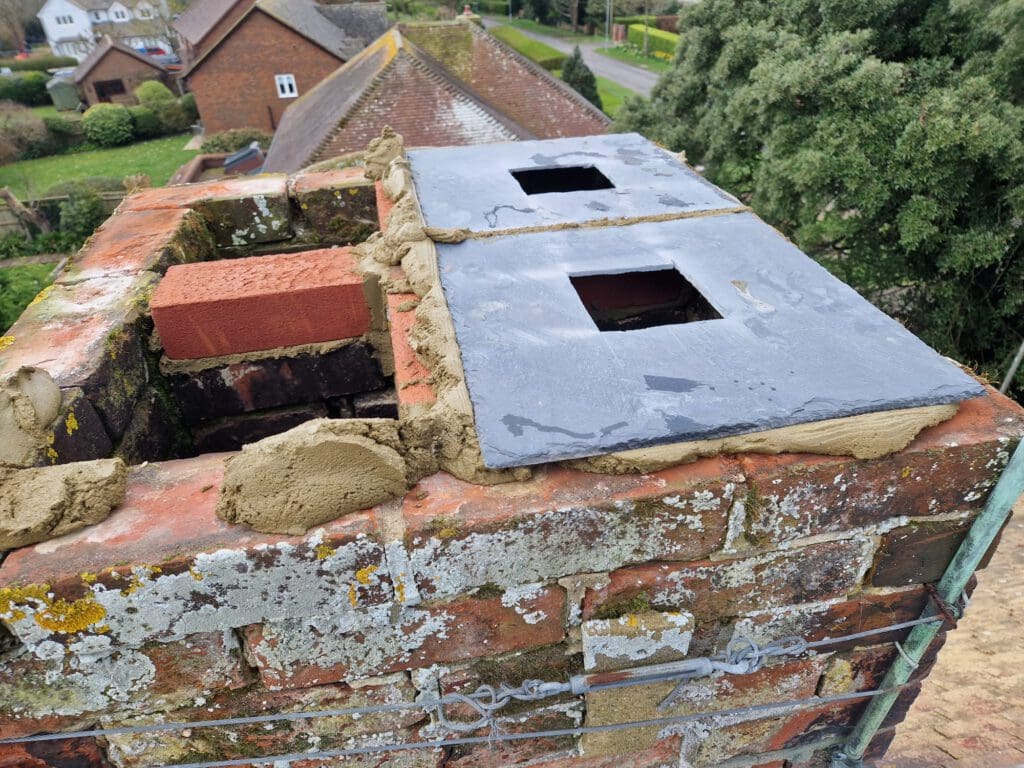 A close-up of a brick chimney with two new slate caps being installed. Fresh mortar surrounds the caps, and the bricks show signs of weathering. The chimney is atop a roof with a view of nearby houses and trees.
