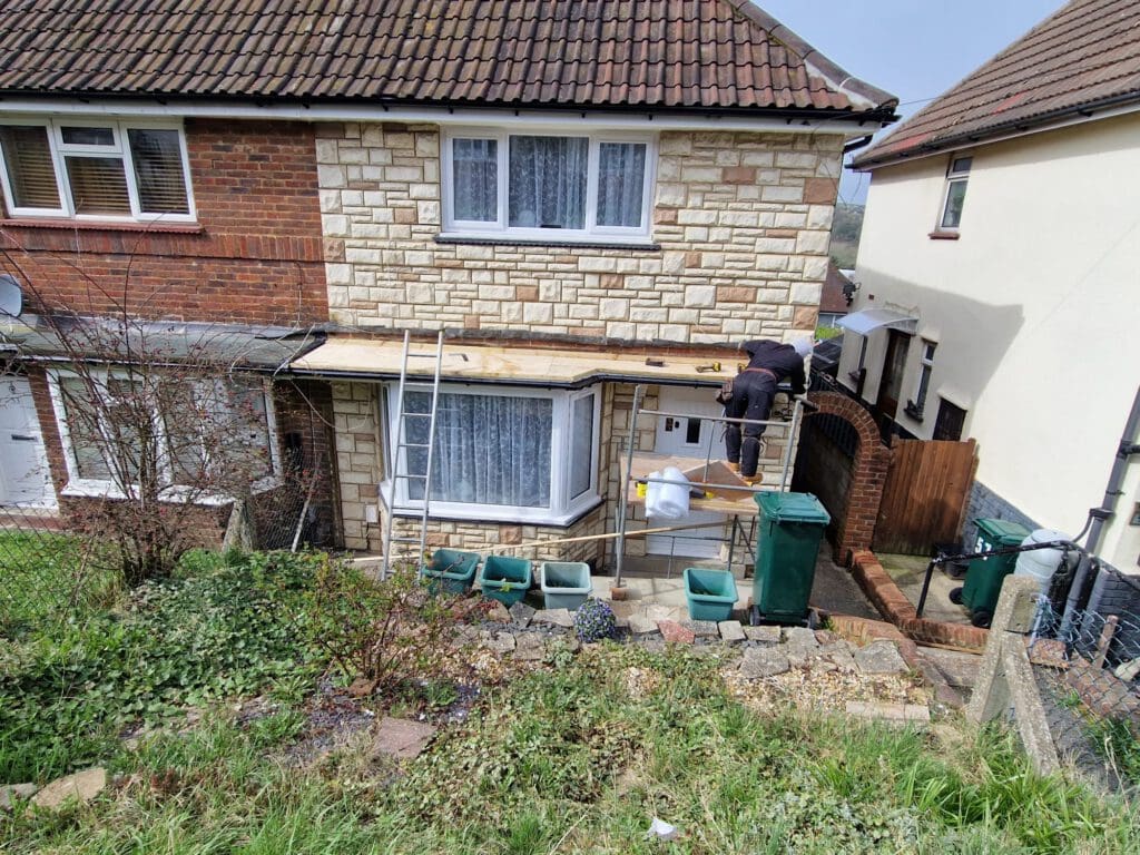 A worker stands on scaffolding performing maintenance on the roof of a two-story house with brick and stone exterior. A ladder leans against the house, and multiple recycling bins are lined up by the entrance.