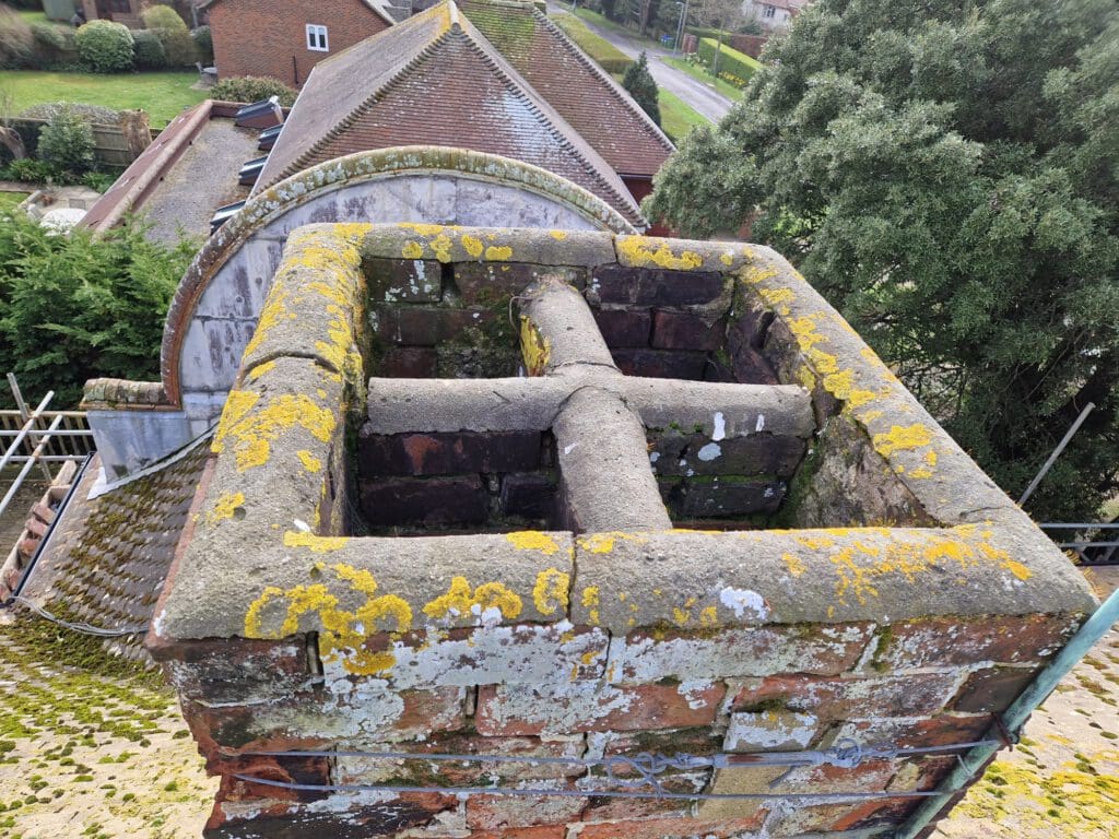 A close-up view of a weathered brick chimney with moss and lichen growth. The background reveals rooftops, a tree, and part of a residential area.