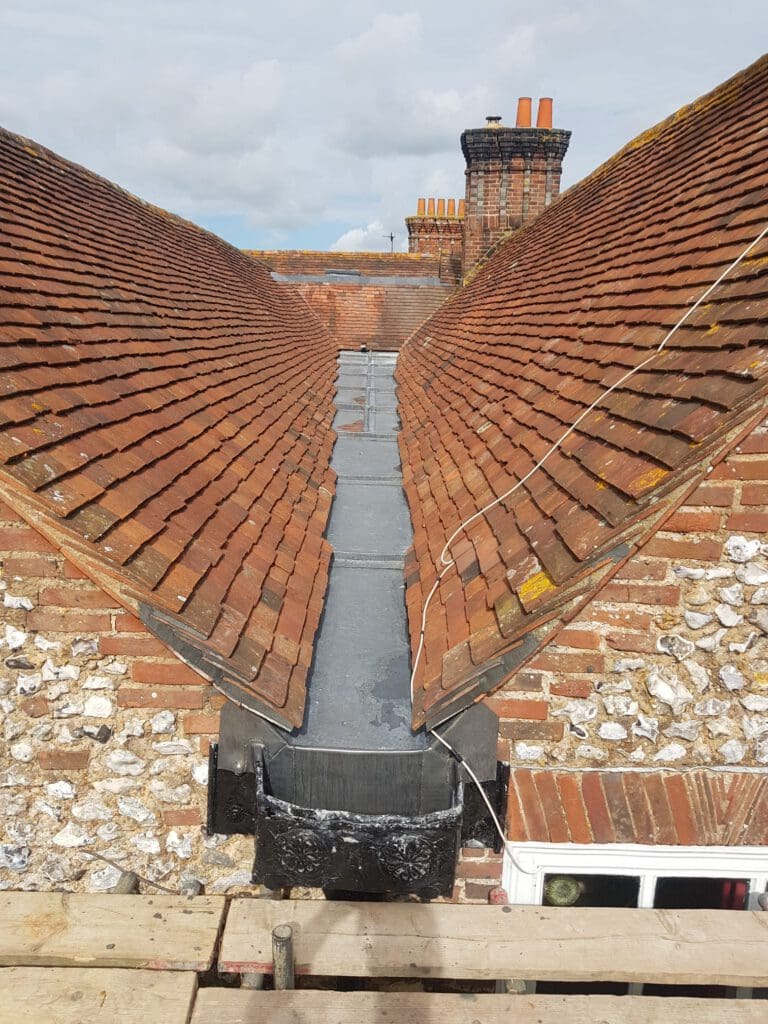 Roof valley with red tiles converging towards a central gutter. There are brick chimneys in the background and a scaffold platform below. Overcast sky in the background.