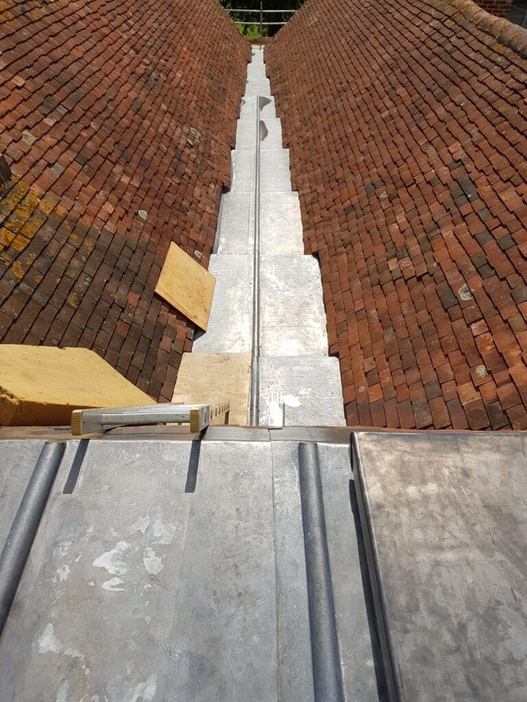 A bird's eye view of a roof with red tiles, featuring a newly installed lead valley drainage system. Wooden boards are placed adjacent to the valley, and sunlight reflects off the metal surface.
