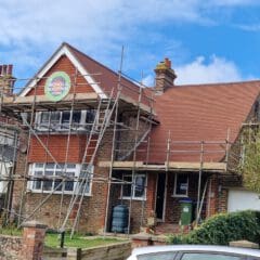 A two-story house with scaffolding surrounding the red-tiled roof for roofing work. A sign on the scaffolding reads "Nichols Roofing." The brick and wood exterior is visible, and trees line the front yard under a clear sky.