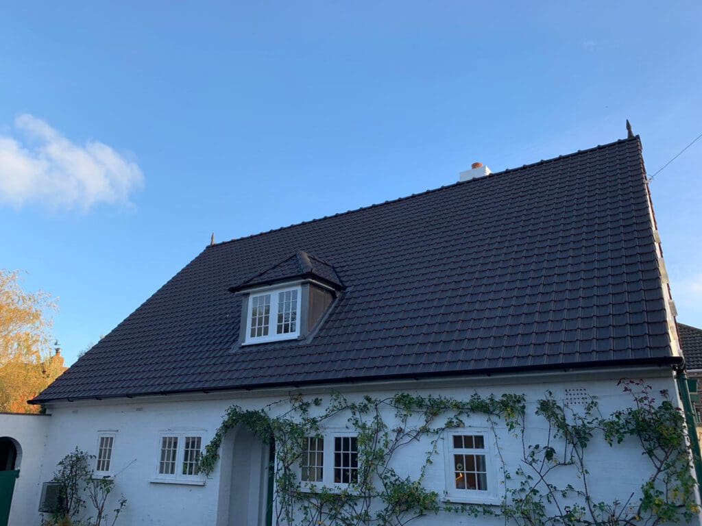 A white house with a steep, dark shingled roof and a small dormer window. The facade features several windows and climbing plants. The sky is clear and blue.