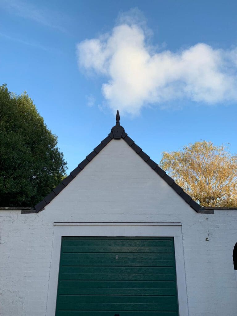 A small white building with a triangular roof, topped by a decorative finial, stands under a clear blue sky. Green trees surround the sides, and a single cloud is visible above, resembling a heart shape. The garage door is painted green.