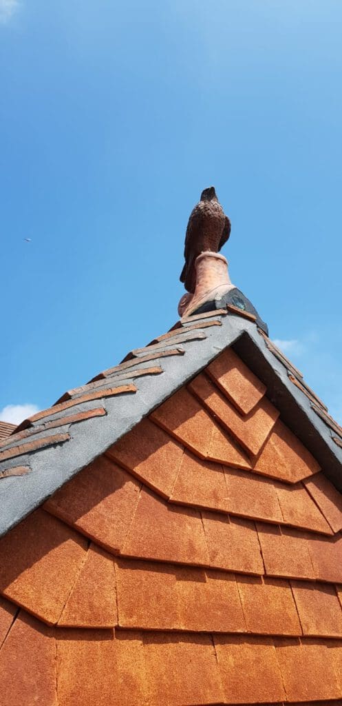 Orange shingled rooftop with a decorative sculpture of a bird perched at the peak against a clear blue sky. A small aircraft is visible in the distance.