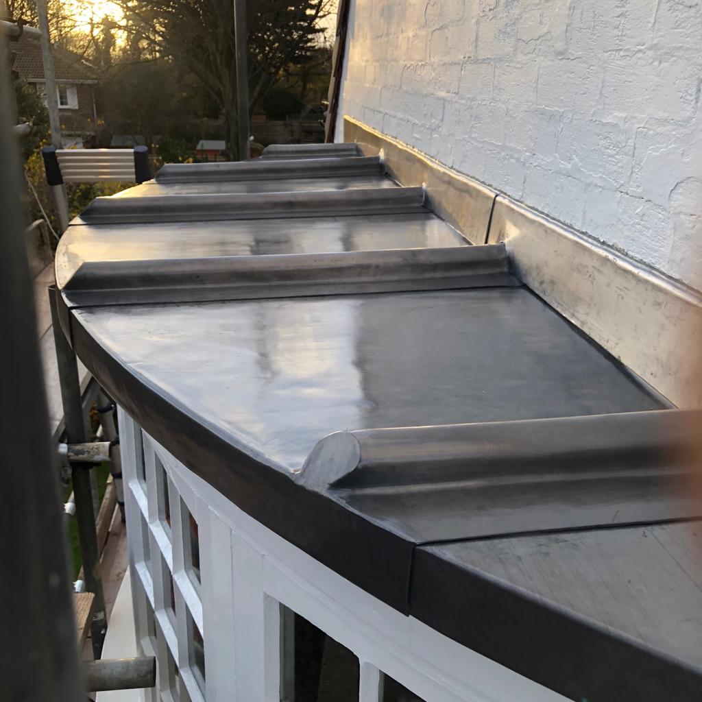 Curved roof with a newly installed flat lead cover, adjacent to a white brick wall. The roof includes ridged sections and overlooks a garden area in the background, visible under the warm glow of the setting sun. A scaffold is visible on the left side.