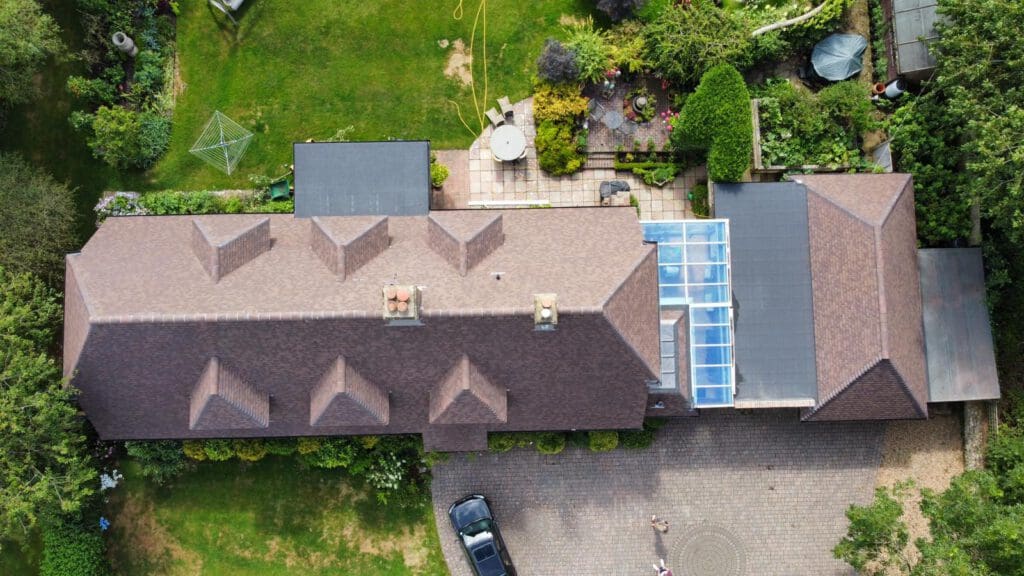 Aerial view of a large house with brown roofs, featuring multiple gables. The property includes a paved driveway, a patio with seating, and lush green gardens surrounding the building. A glass-roofed section extends from one side.